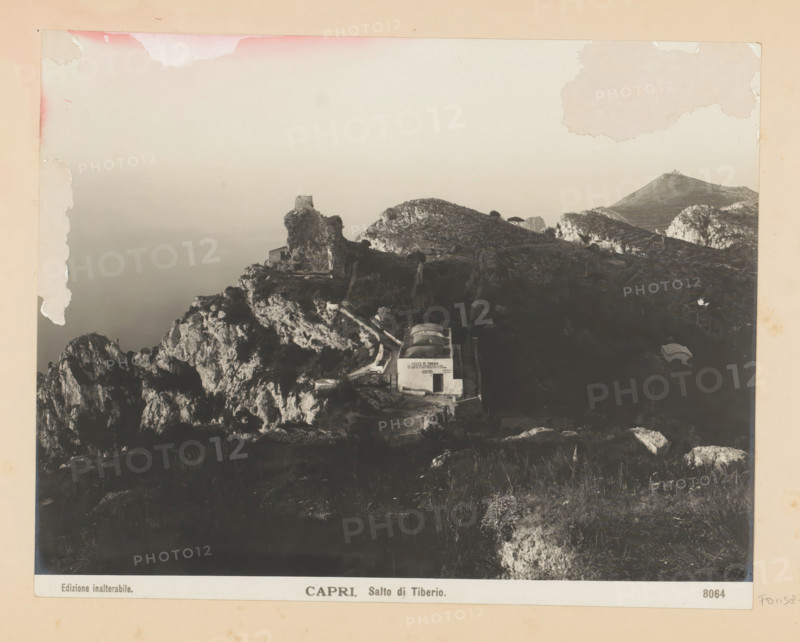 Capri. Salto di Tiberio, View of the Salto di Tiberio in Capri, photograph, Part of Photo album with recordings of sights and works of art in Italy., number: '8064', Capri, c. 1875 - in or before 1907, photographer, paper, gelatin silver print, developing gelatin silver print, height 190 mm x width 242 mm, coastal cliff or crag, Salto di Tiberio, A faded sepia photograph showing a rocky coastal landscape with cliffs and rugged hills descending toward a calm sea, a ruined stone structure perched atop a prominent rock outcrop, a small white building with an arched roof set into the hillside near a winding path, scattered vegetation on the slopes, distant rounded hilltops and rocky formations on the horizon, and areas of discoloration and peeling along the photograph's top and left margins.