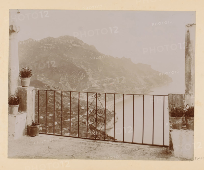 View of a bay (presumably) in Italy, photograph, Part of Photo album with photographs of sights and works of art in Italy., Italy, c. 1875 - in or before 1907, photographer, paper, daylight gelatin silver print, height 195 mm x width 243 mm, gulf, bay, A stone terrace with a metal railing featuring a central decorative scrollwork panel overlooks a coastal cliffside with terraced slopes and a distant shoreline; potted plants sit on short stone pillars at each side of the railing and on the terrace floor, and two tall, weathered columns frame the view.