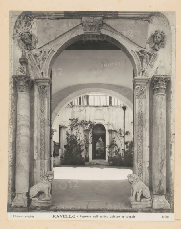 Entrance portal of the episcopal palace of Ravello, Ravello - Ingresso dell' antico palazzo episcopale, photograph, Part of Photo album with photographs of landmarks and works of art in Italy., number: '8199f', Ravello, c. 1875 - in or before 1907, photographer, paper, gelatin silver print, developed gelatin silver print, height 248 mm x width 195 mm, gate, entrance, interior, representation of a building, A stone archway with ornate carved columns frames a courtyard view, showing a series of arches receding into the background and a doorway set into a weathered wall; sculpted angelic faces and decorative reliefs adorn the upper corners and capitals of the columns, while two carved lion statues sit on low plinths at the base of the entrance; potted plants and small trees grow against the walls inside the courtyard, and a narrow railing runs along an upper level above the inner arch.