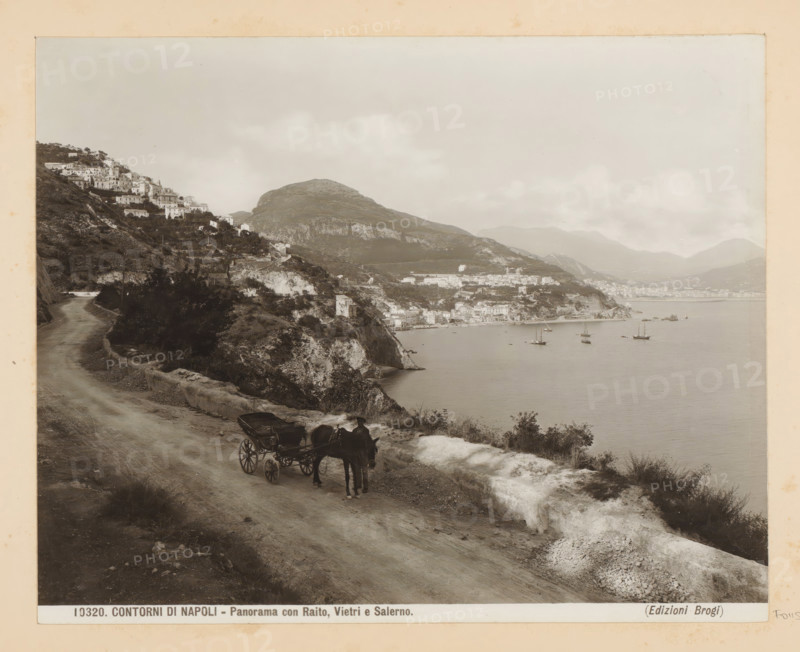 View of Raito, Vietri sul Mare and Salerno, Contorni di Napoli - Panorama con Raito, Vietri e Salerno, photograph, Part of Photo album with recordings of sights and works of art in Italy., number: '10320', photographer: Edizioni Brogi, Salerno (province), c. 1875 - in or before 1907, paper, gelatin silver print, developing gelatin silver print, height 200 mm x width 250 mm, city-view in general, 'veduta', rocky coast, Campagna, Vietri sul Mare, Raito, A coastal scene shows a dirt road running along a hillside with a horse-drawn carriage and driver on the roadside, the horse facing the viewer and the carriage behind it; the road curves toward a village built on terraced slopes with clustered buildings climbing the hillside, and cliffs dropping to a calm bay where several sailing vessels float on the water; distant mountains rise behind the coastal town under a cloudy sky, and vegetation and rocky outcrops line the road and slopes.