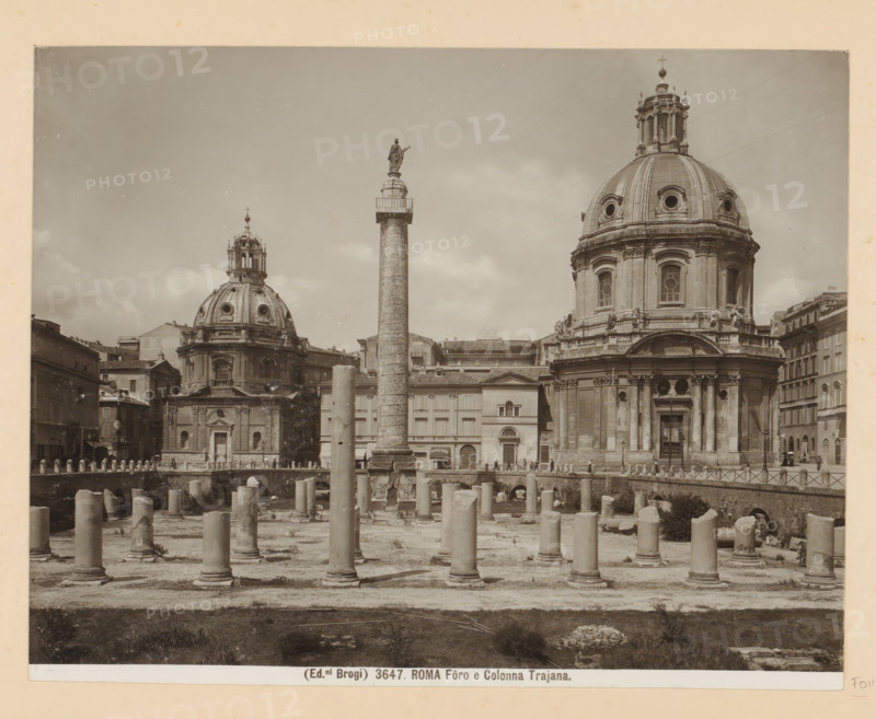Roma Fôro e Colonna Trajana, Roman Forum and Trajan's Column in Rome, photograph, Part of Photo album with recordings of sights and works of art in Italy., number: '3647', photographer: Edizioni Brogi, Forum Romanum, c. 1875 - in or before 1907, Edizioni Brogi, photographer, Rome, Roman Forum, paper, gelatin silver print, developing gelatin silver print, height 200 mm x width 250 mm, landscape with ruins, dome, architecture, triumphal column with helical friezes, Trajan's Column, A sepia photograph showing a wide archaeological site with numerous cylindrical stone columns of varying heights arranged on a dirt surface, a tall ornate column topped with a statue rising near the center, and two large domed classical buildings with detailed façades and circular windows behind the columns; surrounding structures include multi-story buildings and a row of bollards and a railing, and small groups of people are visible near the buildings and along the edge of the ruins.