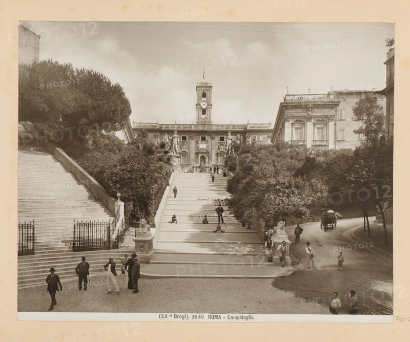 View of the Piazza del Campidoglio in Rome, Roma - Campidoglio, photograph, Part of Photo album with photographs of landmarks and works of art in Italy., number: '3640', photographer: Edizioni Brogi, Rome, c. 1875 - in or before 1907, paper, gelatin silver print, developed gelatin silver print, height 200 mm x width 250 mm, façade (of house or building), staircase, Piazza del Campidoglio, A wide stone staircase flanked by balustrades leads up toward a large ornate building with a central clock tower and statues on pedestals at the top of the steps, trees and dense shrubbery border both sides of the stairs, people in period clothing stand and sit along the stairway and at the base, a horse-drawn carriage is visible on a curved road to the right, stone lion sculptures sit on plinths near the stair entrances, and the scene is framed by additional classical buildings and tall trees under a cloudy sky.