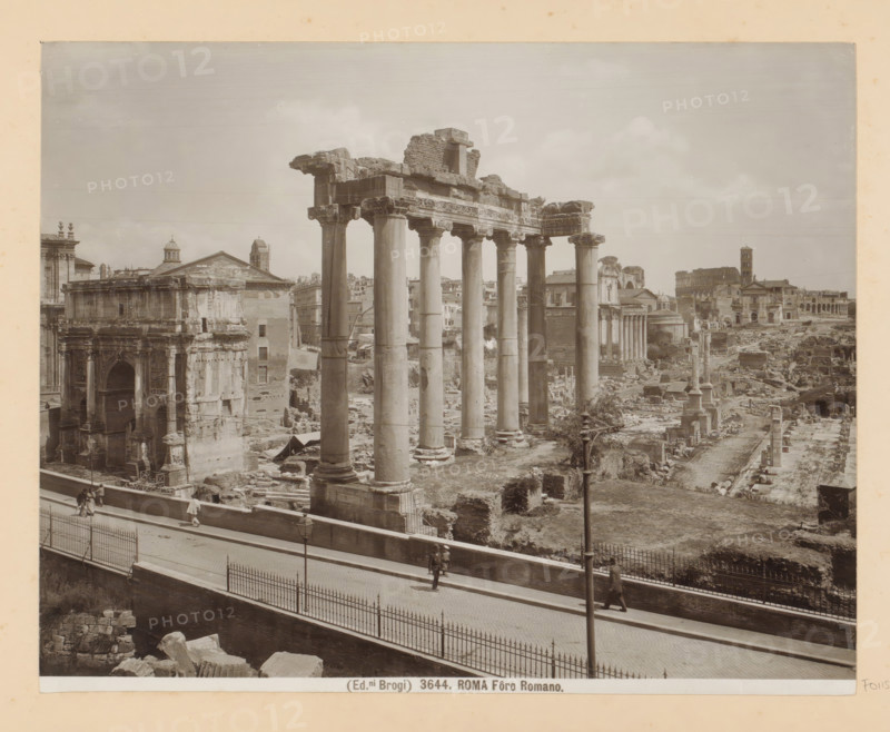 View of the Roman Forum in Rome, Roma Fôro Romano, photograph, Part of Photo album with photographs of sights and works of art in Italy., number: '3644', photographer: Edizioni Brogi, Rome, c. 1875 - in or before 1907, paper, gelatin silver print, developed gelatin silver print, height 200 mm x width 250 mm, landscape with ruins, Forum Romanum, A wide sepia-toned photograph showing a row of tall ancient stone columns with carved capitals supporting a broken entablature, surrounded by scattered ruins and low walls, with additional classical building façades and domes visible behind and beyond them; in the foreground a paved promenade bordered by a metal fence runs parallel to the ruins and shows a few pedestrians walking, while lamp posts and fragments of masonry occupy nearby ground areas.