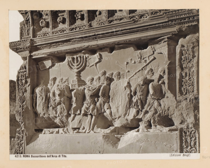 Rome Bas-relief of the Arch of Tito, Bas-relief on the Arch of Titus in Rome, depicting objects taken from the Temple of Jerusalem as booty of war, photograph, Part of Photo album with photographs of sights and artworks in Italy., number: '4213', photographer: Edizioni Brogi, Rome, c. 1875 - in or before 1907, paper, gelatin silver print, height 197 mm x width 250 mm, booty, spoil, sculpture (relief, sculpture), Emperor Titus destroying the temple of Jerusalem, A close-up photograph of a carved stone relief showing a procession of robed figures in profile, some carrying a multi-branched ceremonial object on a pedestal and others bearing long poles and rectangular standards, all rendered in worn, light-colored stone with detailed drapery and facial features; the relief is set within an ornate architectural frame with floral and geometric carvings along the top and sides, a horizontal cornice with layered moldings above the scene, and sculpted decorative panels flanking the figures, with...