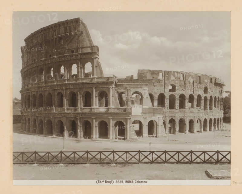 Exterior of the Colosseum in Rome, Roma Colosseo, photograph, Part of Photo album with photographs of landmarks and works of art in Italy., number: '3635', photographer: Edizioni Brogi, Rome, c. 1875 - c. 1907, paper, gelatin silver print, developed gelatin silver print, height 200 mm x width 250 mm, ruin of a building, architecture, Colosseum, A sepia-toned photograph shows the exterior of a large ancient amphitheater with multiple levels of arches and columns, the stone facade weathered and partially ruined, with sections missing and exposed inner walls. The foreground features a low wooden fence with a crisscross pattern running horizontally across the image, and an open paved area between the fence and the building. The sky above is filled with scattered clouds, and small architectural details such as niches, stair openings, and varying arch shapes are visible along the structure.