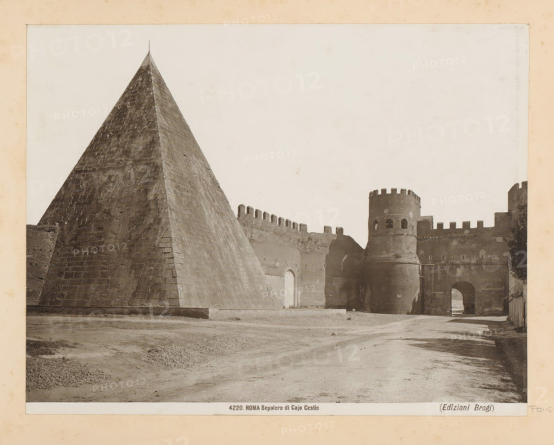 Pyramid of Cestius and the Porta San Paolo in Rome, ROMA Sepolcro di Cajo Cestio, photograph, Part of Photo album with photographs of landmarks and artworks in Italy., number: '4220', photographer: Edizioni Brogi, Rome, c. 1875 - in or before 1907, paper, gelatin silver print, height 193 mm x width 250 mm, pyramid (marking grave), gate, entrance, Porta San Paolo, Pyramid of Cestius, A large stone pyramid with a steep, textured surface occupies the left side, its apex topped by a thin vertical element, while to the right a fortified wall with battlements extends toward a circular tower featuring narrow openings and crenellations, an arched gateway beneath the tower leads into a shadowed passage and a cobbled roadway runs across the foreground with areas of compacted earth and small piles of gravel.