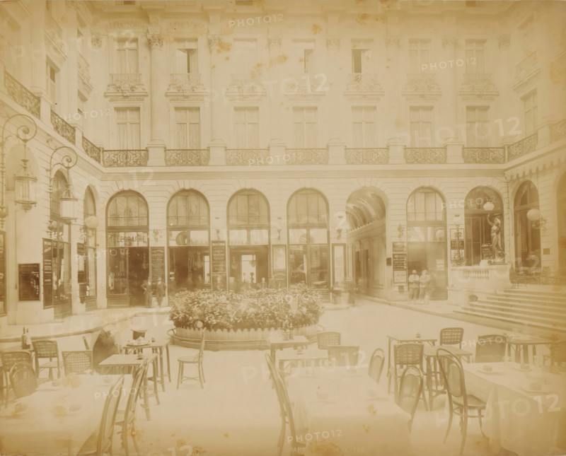 Passage with a terrace in Paris, photograph, Part of Travel Album with photographs of sights in Sweden, Norway, Denmark, Bavaria, Milan and Paris., Paris, c. 1870 - c. 1900, photographer, paper, albumen print, height 221 mm x width 278 mm, pavement, terrace in front of inn or restaurant, advertising, A faded courtyard scene with ornate building facades featuring tall arched doorways and windows, decorative iron railings on a balcony level, sculpted pilasters and ornate cornices, street lamps with curved arms mounted on walls, a covered passage visible through one arch, a raised circular planter filled with dense foliage in the center, café tables and wicker chairs arranged in the foreground with table settings and small objects on them, a few standing figures near the entrances, and a statue on a plinth beside a staircase leading up to another entrance.