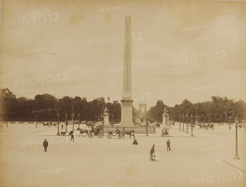 PARIS. - The Place de la Concorde and the Avenue of the Champs-Élysées, View of Place de la Concorde with the Luxor obelisk and the Avenue of the Champs-Élysées in the distance in Paris, photograph, Part of a travel album with photographs of sights in Sweden, Norway, Denmark, Bavaria, Milan, and Paris., number: '1098', photographer: X phot., Paris, c. 1870 - c. 1890, paper, albumen print, height 211 mm x width 275 mm, monument, statue, square, circus, obelisk, needle, Place de la Concorde, Champs-Elysées, A wide open square with a tall slender obelisk rising from a fenced base at its center, flanked by ornate lamp posts and sculpted pedestals, a distant arch visible along the central sightline beyond the obelisk, rows of trees forming a dark band behind the monument, horse-drawn carriages and horses moving along the roadway surrounding the obelisk, several pedestrians scattered across the open paved foreground including a woman with a small child and a few men walking, lamp posts linin...