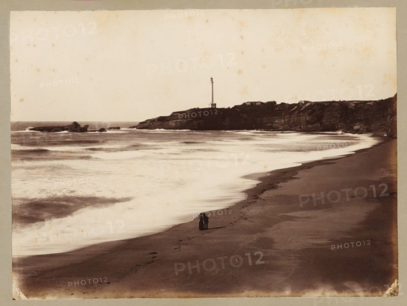 Côte des Fous with lighthouse near Biarritz, album page, photograph, This photo is part of an album., annotation: 'Biarritz, Côte des Fous, ...', 1880 - 1910, photographer, paper, albumen print, height 175 mm x width 234 mm, beach, lighthouse, lightship, Biarritz, A sepia-toned coastal scene showing a sandy beach curving along the shoreline with wet sand reflecting light, gentle waves washing ashore in layered bands, and a rocky headland in the distance topped by a tall, slender lighthouse and a couple of low buildings; a small dark figure stands on the beach near the water's edge, and scattered rocks and a distant rocky outcrop sit offshore beneath a pale sky.