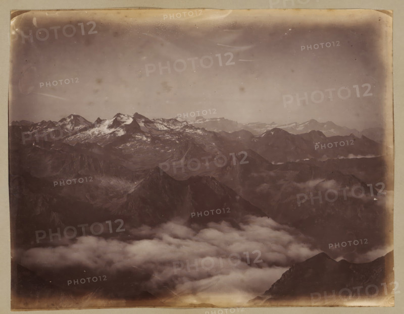 View from the Col d'Aspin, album page, photograph, This photo is part of an album., annotation: 'Vue prise de Col d'Aspin', 1880 - 1910, photographer, paper, albumen print, height 181 mm x width 233 mm, mountains, Occitanie, A sepia-toned photograph showing a panoramic view of rugged mountain peaks with patches of snow on higher summits, layered ridges receding into the distance, and a thick bank of low clouds filling the valleys below, all framed by a worn photographic border with rounded corners and slight discoloration.