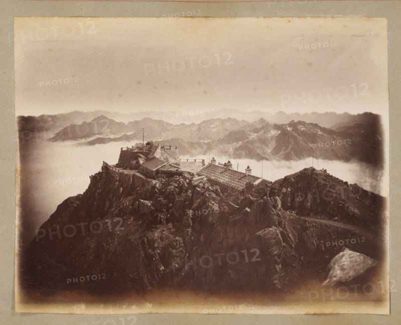 View from Pic du Midi de Bigorre, album page, photograph, This photo is part of an album., annotation: 'Vue prise du Pic du Midi', 1880 - 1910, photographer, paper, albumen print, height 178 mm x width 226 mm, mountains, Midi de Bigorre, Pic du, A sepia-toned photograph showing a rugged rocky mountain peak with a stone building complex and wooden structures perched near the summit, low clouds or mist filling the valleys below, distant jagged mountain ranges visible through the haze, a terrace or platform with railings and flagpoles atop the structure, steep rock faces and boulders in the foreground, and a faded border around the image.