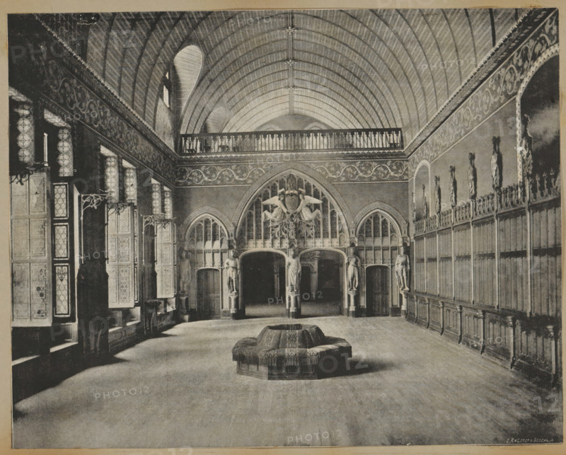 Interior of the castle of Pierrefonds, album page, photomechanical print, This print is part of an album., maker: Angerer & Göschl, C., 1880 - 1900, paper, autotype, height 201 mm x width 248 mm, castle - AA - civic architecture: inside, Pierrefonds, Castle of Pierrefonds, A large vaulted hall with a ribbed arched ceiling and a decorative balcony running along the upper wall is shown, with ornate carved panels and tall narrow windows along one side and a long wooden paneled wall with sculpted figures in niches along the opposite side; at the far end there are arched openings flanked by sculpted statues and an elaborate carved archway and crest, and in the center foreground a circular upholstered seating piece with fringe sits on a broad wooden floor.