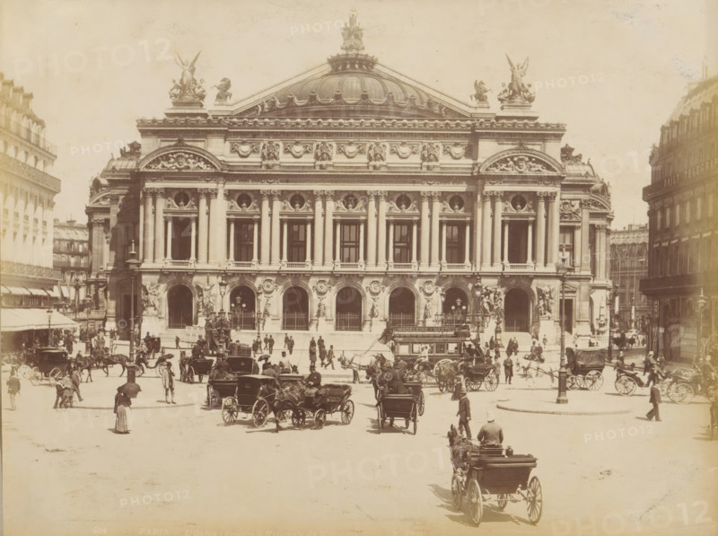 Carriages in front of the Palais Garnier in Paris, album page, photograph, This photo is part of an album., inscription: '604 Paris L'Opéra (Academie Nationale de Musique)', photographer: X phot., 1887 - 1910, X phot., photographer, paper, albumen print, height 208 mm x width 274 mm, historical persons, diligence, omnibus, horse-tram, animal traction, animal-drawn vehicle (carriage etc.), opera house, Paris, Palais Garnier, A wide frontal view of a large ornate stone building with a domed roof and sculptural figures on its roofline, a symmetrical facade with tall columns and arched openings at street level, decorative sculpted reliefs above the columns, and statues flanking the main entrances; in front of the building a busy street scene with many horse-drawn carriages and people walking or standing, some holding umbrellas, and a larger carriage approaching in the foreground with a driver seated at the front; surrounding the central building are multi-story urban buildings on either si...