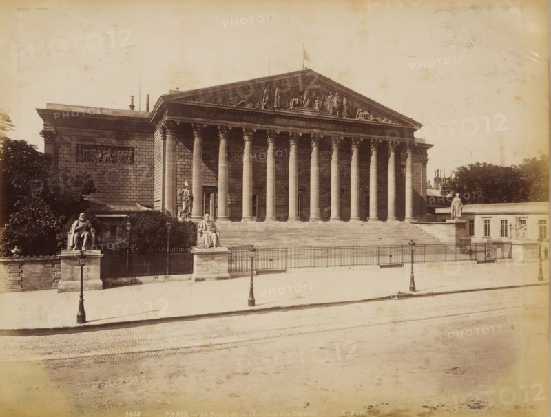 Palais Bourbon in Paris, album page, photograph, This photo is part of an album., inscription: '1488 Paris _ Le Palais de la Chambre des Députés', photographer: height 208 mm x width 272 mm, front facade, civic architecture, edifices, dwellings, Paris, Palais Bourbon, A large neoclassical stone building with a wide flight of steps leading to a portico of tall fluted columns topped by a triangular pediment with sculpted figures, flanked by blocky wings with bas-relief panels; stone statues on pedestals stand at the corners and near the stairs, and ornate street lamps line the fenced area in front; trees and shrubbery appear behind and beside the building, and a paved street and sidewalk occupy the foreground.