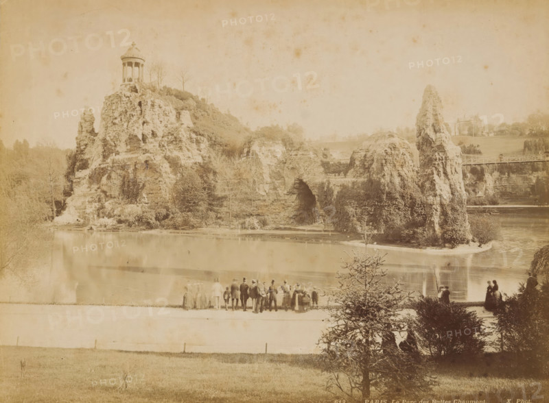 Spectators at Parc des Buttes-Chaumont in Paris, album page, photograph, This photo is part of an album., inscription: '613 Paris _ Le Parc des Buttes-Chaumont', photographer: X phot., 1887 - 1910, paper, albumen print, height 204 mm x width 273 mm, historical persons, public gardens, park, Paris, Parc des Buttes-Chaumont, A sepia-toned landscape showing a rocky hill formation rising from a body of water, with a round columned pavilion perched atop the highest rock and bare trees nearby. The rocky slope has textured cliffs, cavities, and vegetation clinging to its sides, and a tall narrow rock stack juts upward near a bridge that spans a gap to a grassy area with scattered trees and a distant building. In the foreground a group of people stand along the water's edge facing the rocks, some in long coats and hats and a few women in long dresses, while others are seated or walking near bushes and a small conifer, and reflections of the shoreline appear on the water.