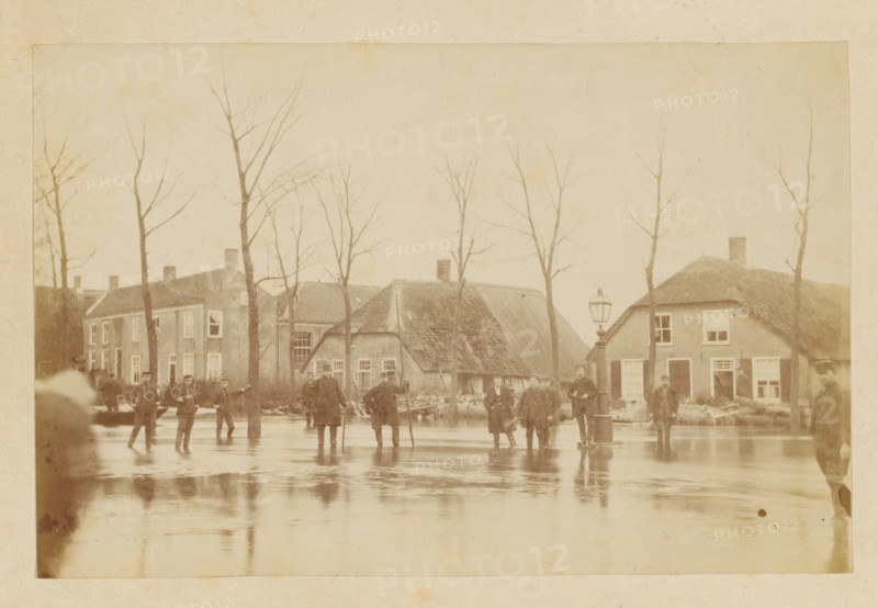 Flooding Nieuwkuyk 1916, Flood in Nieuwkuijk, photograph, photographer: attributed to Albert Greiner, Amsterdam, 1880 - 1881, Attributed to, pasteboard, paper, albumen print, albumendruk, height 100 mm x width 153 mm, height 210 mm x width 256 mm, the year 1916 should be 1880, village, flood, overvloed, Nieuwkuijk, Period, 1880-12-29 - 1880-12-30, A sepia-toned street scene showing a row of houses with thatched and tiled roofs behind a line of leafless trees, a group of people standing and walking on a reflective wet surface in front of the houses, a tall street lamp near the center right, several figures wearing coats and hats, and the buildings' windows and doors visible along the street.