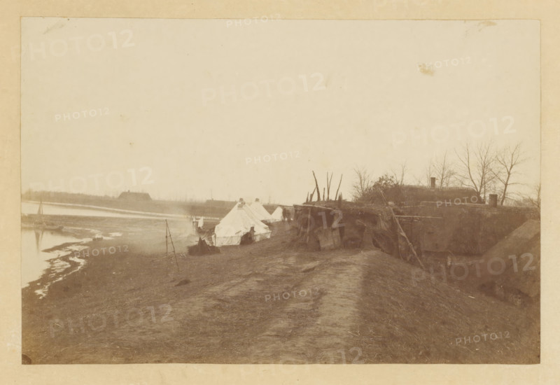 Flood in Nieuwkuijk, Flood Nieuwkuyk N Br. 1916, photograph, title: 'Overstroming Nieuwkuyk N Br. 1916', photographer: attributed to Albert Greiner, Amsterdam, 1880 - 1881, Attributed to, pasteboard, paper, albumen print, albumendruk, height 105 mm x width 155 mm, height 210 mm x width 262 mm, Date 1916 is incorrect, should be 1880, village, flood, overstroming, Nieuwkuijk, Period, 1880-12-29 - 1880-12-30, A sepia-toned scene showing a riverside encampment with several canvas tents pitched along a dirt embankment, a narrow path running between the tents and the camera, and makeshift wooden structures and poles nearby; water and a muddy shoreline appear to the left with a small boat pulled up near the bank, and low buildings and leafless trees are visible in the distant background on the right.