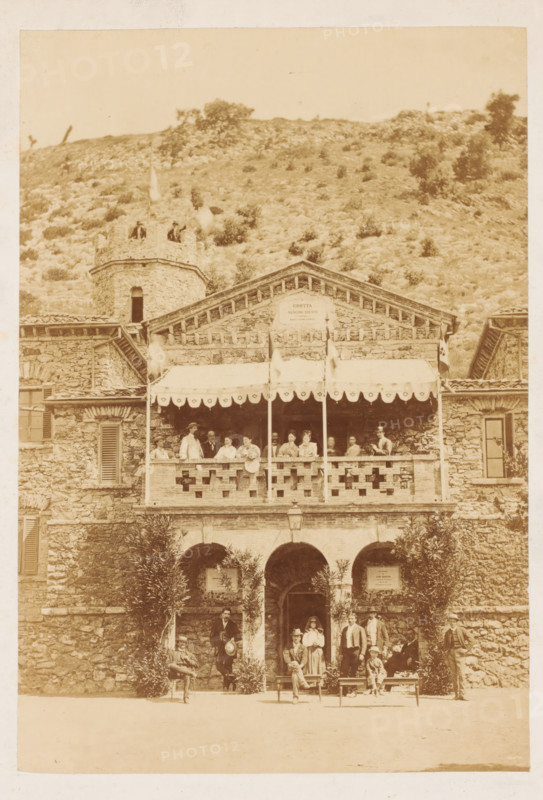 Entrance to the Grotta Giusti in Pistoia, photograph, photographer: Scevola Ghelli, Pistoia, 1860 - 1900, Tuscany, pasteboard, paper, albumen print, albumendruk, height 201 mm x width 139 mm, height 250 mm x width 224 mm, watering-place, spa, Grotta Giusti, A sepia-toned photograph showing a stone building with a balcony and an awning, the balcony edge decorated with a pattern of openings and supported by posts, a group of people standing and sitting along the balcony railing, and additional people posed on the ground level in front of arched doorways framed by foliage; the building has shuttered windows, a small round tower rising to one side with people visible on its top, and a hillside covered in sparse vegetation visible behind the structure.