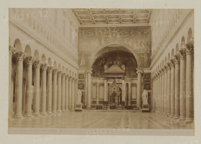 Basilica di S. Paolo furoi le mura, Interior of St. Paul outside the Walls in Rome, Italy, Roma, photograph, number: '...', photographer: Fratelli Alinari, Rome, 1852 - 1900, pasteboard, paper, albumen print, height 97 mm x width 144 mm, height 168 mm x width 223 mm, interior of church, Saint Paul Outside the Walls, A sepia-toned interior view showing a long central nave flanked by rows of tall columns with ornate capitals, arched openings above the columns on both sides, a patterned coffered ceiling, a polished floor reflecting the columns, sculpted statues on pedestals near the front, and a decorated apse with a large semicircular mosaic or painting featuring a central haloed figure and surrounding figures.