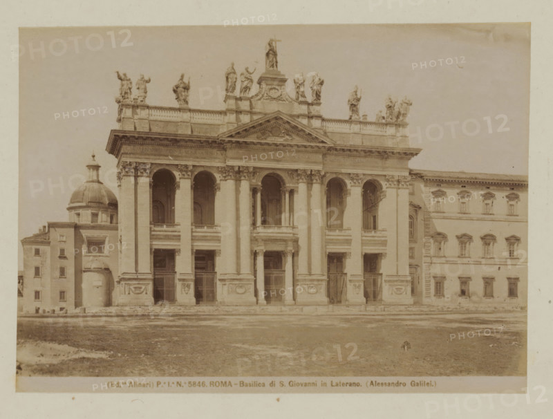 West facade of St. John Lateran in Rome, Italy, Basilica di S. Giovanni in Laterano (Alessandro Galilei), Roma, photograph, number: 'Po. Io. No. 5846.', photographer: Fratelli Alinari, Rome, 1852 - 1900, pasteboard, paper, albumen print, height 101 mm x width 141 mm, height 168 mm x width 224 mm, parts of church exterior and annexes: west facade, Sint-Jan Lateranen, A sepia photograph of a large classical building façade featuring tall columns and arched openings, with a triangular pediment above the central section and sculpted statues lined along the roofline; to the left a domed structure and adjacent rectangular building with small windows; to the right a multi-story building with regularly spaced windows and decorative window frames; the foreground shows an open paved and slightly grassy area leading up to the building steps, and the sky above is clear and pale.