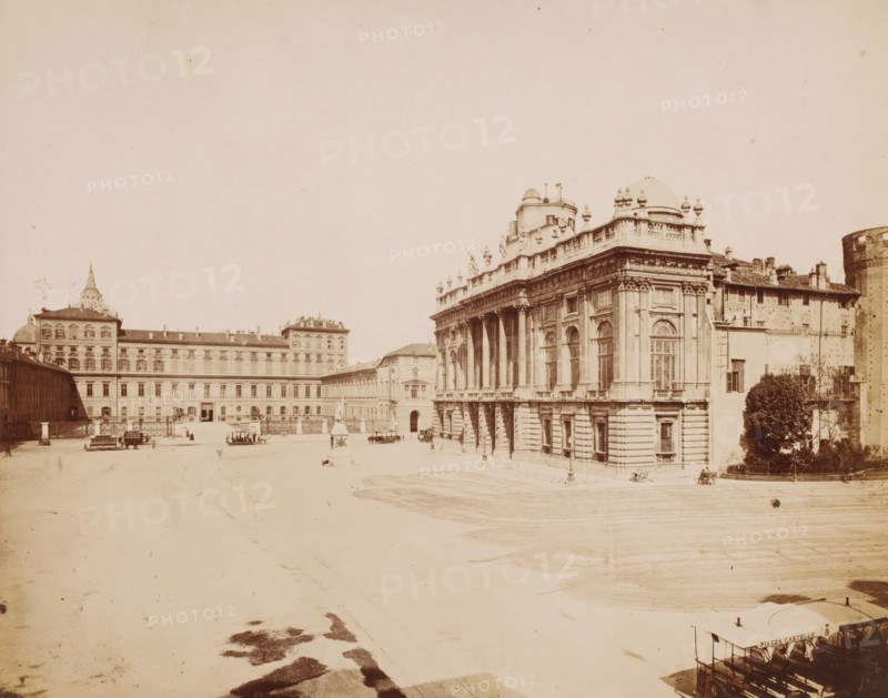 View of the Piazza Castello in Turin, Italy, Piazza Castella, Torino, photograph, address: 'VIA GERRAIA CORSO SICCARDI, TORINO', photographer: Fotografia Subalpina, Turin, 1899 - 1910, Piedmont, pasteboard, paper, albumen print, height 260 mm x width 316 mm, square, circus, exterior, representation of a building, Piazza Castello (Turin), A wide open paved square bordered by large classical buildings with ornate facades, tall arched windows, columns, balustrades, and sculptural details, one prominent building with domes and ornamental statues along its roofline at the right, an extended multi-story building stretching across the background to the left with a decorative roof element, scattered small horse-drawn carriages and benches positioned across the square, a lone tree and shrubbery near the right edge beside a simpler building, a cylindrical tower partially visible at the far right, and an overall pale, sepia-toned appearance.