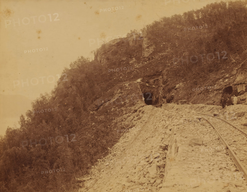 View of the Luleå - Narvik railway line, on the right some railway workers, photograph, Sweden, 1888 - 1900, photographer, pasteboard, paper, albumen print, height 195 mm x width 253 mm, height 204 mm x width 259 mm, railway, train, Nordland, Luleå, Narvik, A sepia-toned landscape showing a rocky hillside with sparse trees and vegetation, a dark tunnel opening carved into the rock near the center, rough gravel and debris leading toward the tunnel, narrow-gauge rails running along the ground toward the right side, a small group of people standing near the tunnel entrance and beside another darker opening with a wooden structure, scattered rocks and wooden planks along the path, and an open pale sky occupying the upper left portion of the image.