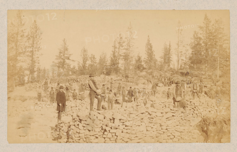 Group of men, possibly miners in Northern Sweden between Luleå and Narvik, photograph, Sweden, 1891 - 1900, photographer, pasteboard, paper, albumen print, height 121 mm x width 203 mm, height 240 mm x width 333 mm, miner, A wide sepia photograph shows a rocky outdoor scene with a group of people standing among piles of irregular stones and low stacked stone walls, tall evergreen trees rising in the background, several men wearing hats and work clothes scattered across the rocky ground, a few individuals standing on higher stone piles, one man in the foreground facing the camera holding an object, another man nearby standing with a tool, and additional figures dispersed along terraces of stone with trees and rough terrain beyond.