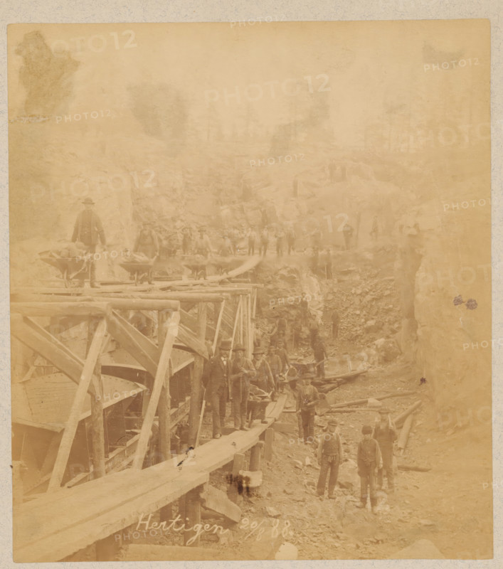 Works in the Hertigen mine near Malmberget, Sweden, photograph, Malmberget, 1888-08-20, photographer, pasteboard, paper, albumen print, albumendruk, height 182 mm x width 164 mm, height 332 mm x width 242 mm, quarry (stone), Hertigen, A faded sepia photograph showing a group of men standing and walking on a rough, rocky worksite with wooden scaffolding and planks forming a raised walkway along one side; some men stand on the walkway while others are on the uneven ground below, many wearing hats and work clothes, and a few appear to be dressed more formally; large wooden beams and structural supports are visible in the foreground, with piles of rocks and timbers scattered throughout the scene and a steep rocky face rising at the back where additional figures stand on ledges.