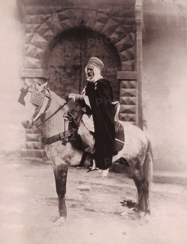 Officer on horseback, probably in Algiers, Officer Indischene, photomechanical print, inscription: 'foto Leroux Alger', photographer: Alexandre Leroux, Algiers, cliché maker: anonymous, 1880 - 1900, North Africa, pasteboard, paper, collotype, photoprint, height 317 mm x width 247 mm, riding a horse, ass, or mule, rider, horseman, A sepia-toned photograph shows a man seated on a horse in front of a large stone archway with a textured pattern. The man wears a wrapped head covering and a long dark cloak draped over lighter garments, and he has a visible beard. The horse is outfitted with ornate decorative tack including a patterned headpiece and a decorated breast collar, and the saddle area shows embroidered fabric and straps. The ground beneath the horse appears uneven and there are faint shadows and marks on the stone surface and surrounding wall.