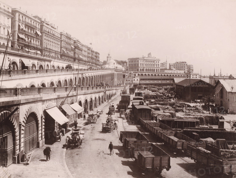 Les Quais à Alger, Quays in Algiers, photomechanical print, number: '1563', photographer: Alexandre Leroux, Algiers, cliché maker: anonymous, c. 1875 - c. 1900, North Africa, pasteboard, paper, collotype, photoprint, height 247 mm x width 318 mm, warehouse (, harbour), A wide city quay scene with a long colonnaded building on the left featuring repeated arched openings and balconies above, rows of awnings and railings, and multi-story buildings rising behind it; a broad street runs alongside the building filled with horse-drawn wagons, carts, and people walking or standing; to the right an open yard contains many covered loads, wooden crates and rail wagons, and several low industrial sheds and warehouses with pitched roofs; in the distance more ornate multi-story buildings and a domed structure are visible against a pale sky.