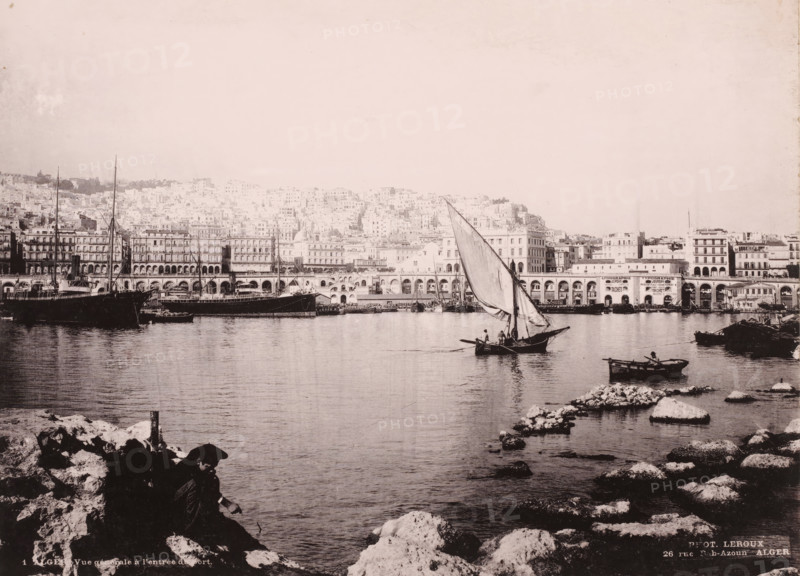 Alger, vue générale à l'entrée du port, View of the port of Algiers, photomechanical print, number: '1', address: '26 rue Bab-Azoun ALGER', photographer: Alexandre Leroux, Algiers, cliché maker: anonymous, c. 1875 - c. 1900, Alexandre Leroux, photographer, cliché maker, North Africa, pasteboard, paper, collotype, photoprint, height 247 mm x width 318 mm, harbour, A wide waterfront scene with calm water in the foreground and a rocky shoreline at the bottom edges, a small sailboat with a large triangular sail and a few people aboard positioned near the center right, a smaller rowboat with a seated person closer to the right edge, several larger moored ships and boats along the left and middle distances, a long quay with arched openings and multi-story buildings behind it stretching across the middle of the image, a densely built hillside of light-colored buildings rising behind the quay, and a person sitting on the rocks in shadow in the lower left foreground.
