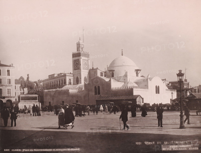 Alger, Place du Gouvernement et mosquée El-Djedid, Djama?a el Djedid mosque in Algiers, photomechanical print, address: '26 rue Bab-Azoun ALGER', number: '1001', photographer: Alexandre Leroux, Algiers, cliché maker: anonymous, c. 1875 - c. 1900, North Africa, pasteboard, paper, collotype, photoprint, height 246 mm x width 318 mm, temple, shrine, Islam, Mohammedanism, Djama?a el Djedid, A wide city square scene with many people walking and standing, some wearing long robes and head coverings, others in coats and hats, and a few animals and carts along the edges; a large mosque-like white building with domes and crenellated walls dominates the middle, accompanied by a tall clock tower with decorative panels and a flagpole, and adjacent low buildings with balconies; a prominent equestrian statue on a pedestal stands to the left, and ornamental street lamps and a small wooden structure are visible near the foreground, with long shadows cast across the open paved area under a pale sky.