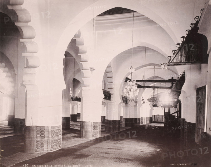 Interieur de la Mosquee El-Kebir-Alger, Interior of the Djama'a al-Kebir in Algiers, photomechanical print, number: '487', photographer: Alexandre Leroux, Algiers, cliché maker: anonymous, c. 1875 - c. 1900, Alexandre Leroux, photographer, cliché maker, North Africa, pasteboard, paper, collotype, photoprint, height 247 mm x width 318 mm, temple, shrine, Islam, Mohammedanism, Djama'a al-Kebir, A large interior space with a series of tall, rounded arches receding into the distance, each arch supported by thick columns whose lower portions are wrapped in patterned coverings, textured floor coverings laid across the ground, ornate hanging chandeliers suspended from the ceiling, pairs of decorative wooden or metal balcony-like structures attached to some arches, carved or scalloped arch details at the tops of several supports, a darker raised platform or structure along one side with mounted lights, high plain walls and vaulted ceilings, and a door opening visible at the far right.