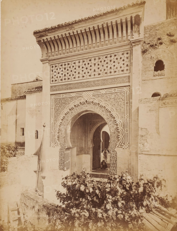 Mosquée Boumédine Tlemcen, Entrance of the Sidi Boumédienne mosque in Tlemcen, Algeria, photograph, inscription: 'Morocco', number: '234', Tlemcen, 1860 - 1900, photographer, North Africa, pasteboard, paper, albumen print, height 317 mm x width 247 mm, temple, shrine, Islam, Mohammedanism, Sidi Boumédienne mosque, A sepia-toned photograph of an ornate architectural façade featuring a large horseshoe-shaped arch with intricate geometric and floral tilework, framed by decorative panels and a carved band above. The upper section shows a lattice-like screen with repeating star patterns beneath a projecting cornice supported by a row of carved brackets. Flanking walls of plain plaster and brick rise on either side, one with small arched openings. Through the archway a passage and additional arches are visible, and two figures stand and sit near the entrance. In the foreground dense foliage and a low stone wall occupy the lower portion of the image.