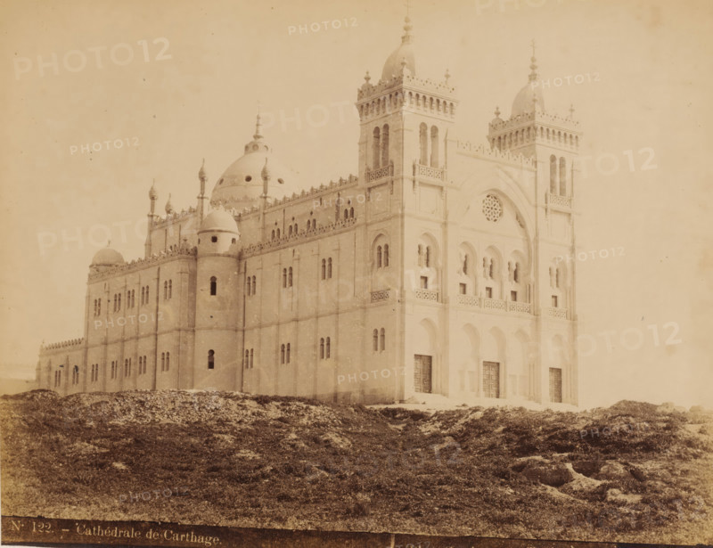 View of the Cathedral of Saint-Louis on the Byrsa in Tunisia, Cathédrale de Carthage, photograph, number: 'N. 122.', photographer: J. André Garrigues, Byrsa, 1884 - c. 1900, North Africa, pasteboard, paper, albumen print, height 247 mm x width 317 mm, church (exterior), Saint-Louis Cathedral, A large pale building with ornate towers and domes sits on a rocky, grassy foreground; the facade shows arched doorways and windows, decorative balustrades and carved details, narrow vertical openings in the towers, a circular rose window set within a recessed arch, and small pinnacles along the roofline.