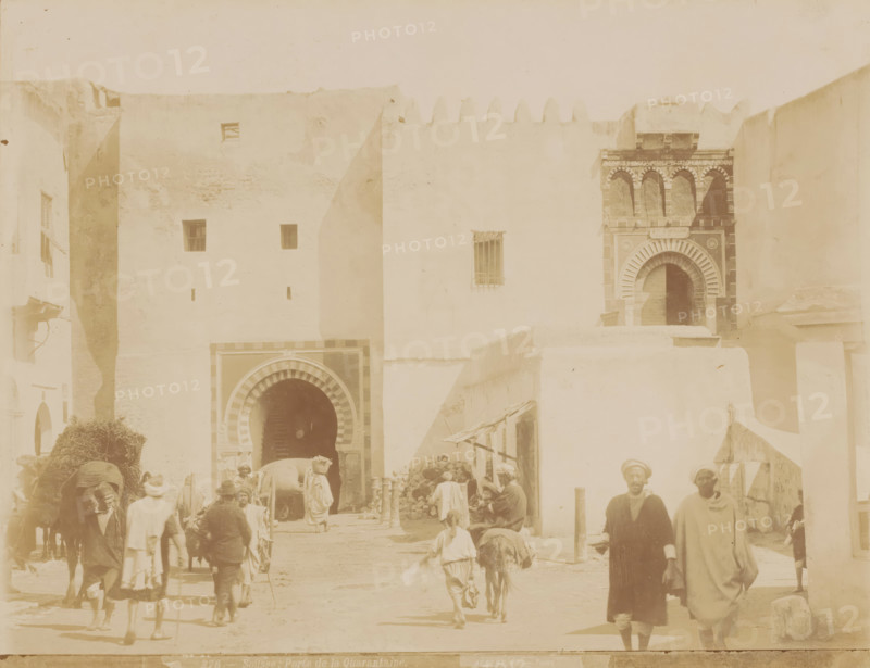 Cityscape in Sousse, Sousse, Porte de la Quarantaine, photograph, number: '276', photographer: Albert Goupil, 1860 - 1900, North Africa, pasteboard, paper, albumen print, albumendruk, height 247 mm x width 317 mm, passage, A pale street scene framed by tall, light-colored walls with small square windows and an ornate arched doorway set into a darker patterned façade, showing several people in loose garments and head coverings walking or standing in the foreground, animals including a donkey or mule and a larger pack animal near the central arch, stacked bundles or hay and a small pile of stones beside a low wall, a second arched entrance partially visible with striped archway decoration, and simple wooden awnings and poles casting soft shadows on the ground.