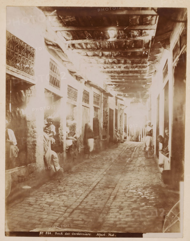 Souk des Cordonniers, Shoemakers in a souk, presumably in the Medina of Tunis, photograph, number: 'No 260.', photographer: Albert Goupil, Medina of Tunis, 1860 - 1900, North Africa, Tunis, pasteboard, paper, albumen print, height 317 mm x width 247 mm, shoe-maker, cobbler, A narrow covered alley with a cobblestone path running down its center is lined by walls with small recessed shop openings and decorative metal grilles, a wooden beam ceiling overhead showing exposed planks and support timbers, and several figures wearing loose garments and headwraps seated or standing in the doorways and entrances along both sides; light filters in from the far end creating bright highlights and soft shadows, and worn textures are visible on the stone and plaster surfaces.