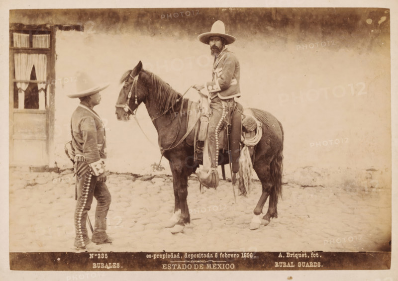 Rurales, Rural Guards, Two Mexican men in traditional clothing, one of them on horseback, Estado de Mexico, photograph, copyright notice: 'es-propriedad, depositada 6 febrero 1896.', number: 'N. 235', photographer: Abel Briquet, Mexico, 1896, North America, pasteboard, paper, albumen print, height 133 mm x width 194 mm, height 160 mm x width 210 mm, adult man, horse, A sepia photograph shows two men and a horse on a cobblestone surface in front of a pale wall with a window partially draped with curtains; one man stands facing the horse wearing a wide-brimmed hat and a decorated jacket and trousers, the other sits astride the horse wearing a similar wide-brimmed hat, a jacket with decorative trim, and a beard, holding the reins; the horse is saddled with ropes, blankets, and gear hanging from the saddle, and both men's clothing and the horse's tack feature rows of round metallic elements.