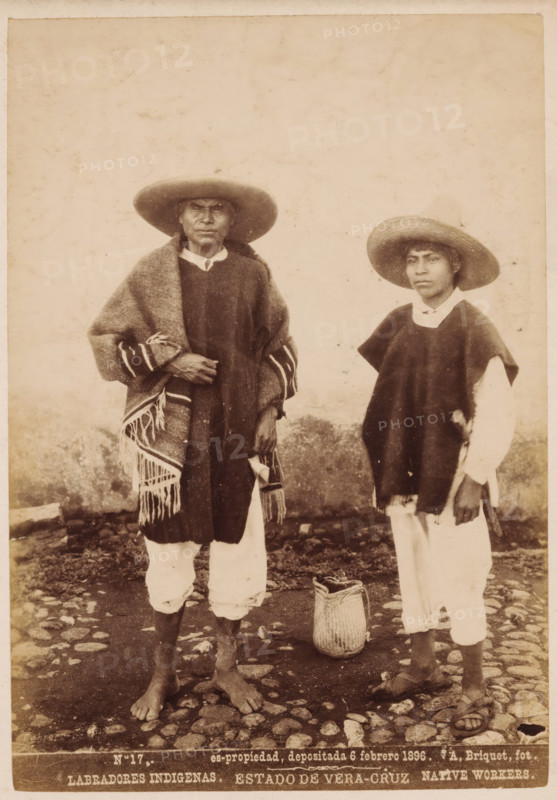 Labradores Indigenas, Native workers, Double portrait of two unknown inhabitants of Mexico, Estado de Vera-Cruz, photograph, copyright notice: 'es-propriedad, depositada 6 febrero 1896.', number: 'N. 17.', photographer: Abel Briquet, Veracruz, 1896, North America, pasteboard, paper, albumen print, height 194 mm x width 133 mm, height 210 mm x width 160 mm, adult man, An aged sepia photograph shows two males standing on a cobblestone ground in front of a plain wall; both wear wide-brimmed hats and loose garments layered over light-colored trousers, with the figure on the left draped in a fringed woven blanket and standing barefoot, the figure on the right wearing a dark poncho over a light shirt and sandals, and a woven basket placed on the ground between them.