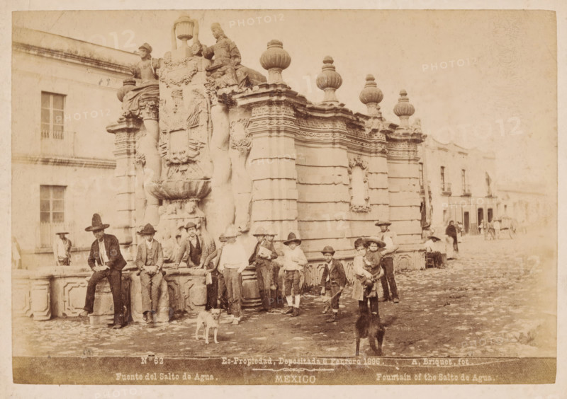 Fuente del Salto de Agua, Fountain of the Salto de Agua, View of the Salto del Agua fountain in Mexico City, Mexico, photograph, A group of men and children sit on the edge of the fountain., copyright notice: 'Es-Propriedad, Depositada 6 Febrero 1896.', number: 'N. 62', photographer: Abel Briquet, Mexico City, 1896, North America, pasteboard, paper, albumen print, height 132 mm x width 193 mm, height 160 mm x width 210 mm, ornamental fountain, Salto del Agua, A sepia-toned street scene shows an ornate stone fountain with sculpted figures, decorative urns, and carved reliefs, positioned against a building facade. Several boys and young men wearing hats and period clothing are gathered around and sitting on the fountain, some holding small objects or leaning on the stone ledge. Two dogs stand on the cobblestone street in front of the group. In the background, additional buildings with windows and balconies line the street and a few distant figures and a horse-drawn cart are visible. The ...