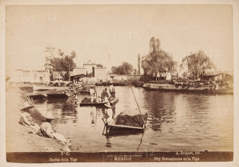 Garita de la Viga, City Customhouse at la Viga, Pramen in the La Viga Canal in Mexico City, Mexico, photograph, number: 'N. 31', photographer: Abel Briquet, Mexico City, 1865 - 1910, North America, pasteboard, paper, albumen print, height 133 mm x width 194 mm, height 160 mm x width 210 mm, canals, waters (in city), city-gate, La Viga canal, A sepia-toned waterfront scene shows a calm body of water with several small flat boats, some loaded with sacks and vegetation, and people seated or standing in them wearing wide-brimmed hats and loose clothing; a few figures sit or recline on the grassy bank in similar attire. Along the opposite shore there are low buildings, a decorative arched gateway, trees with varying heights and foliage, and a narrow pier or dock where boats are moored. Reflections of the boats and people ripple on the water surface and a long pole extends from one vessel into the foreground. The overall image has a faded, aged appearance with soft contrasts and visible pape...