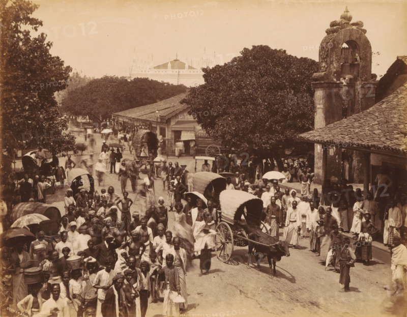 View of the Main Street, Colombo, Sri Lanka, photograph, signature: 'Scowen & Co.', title: 'Main Street, Colombo', photographer: Charles T. Scowen & Co., 1876 - 1893, South Asia, paper, albumen print, height 215 mm x width 277 mm, height 347 mm x width 400 mm, street, A crowded street scene with many people walking and standing, some carrying baskets or bundles and others holding umbrellas, several horse-drawn carts and covered wagons among the crowd, a large tree shading part of the street near a row of single-story buildings with tiled roofs, a stone bell tower with decorative carving and visible bells, a distant ornate building partially visible above the trees, and a variety of clothing styles visible on the people.