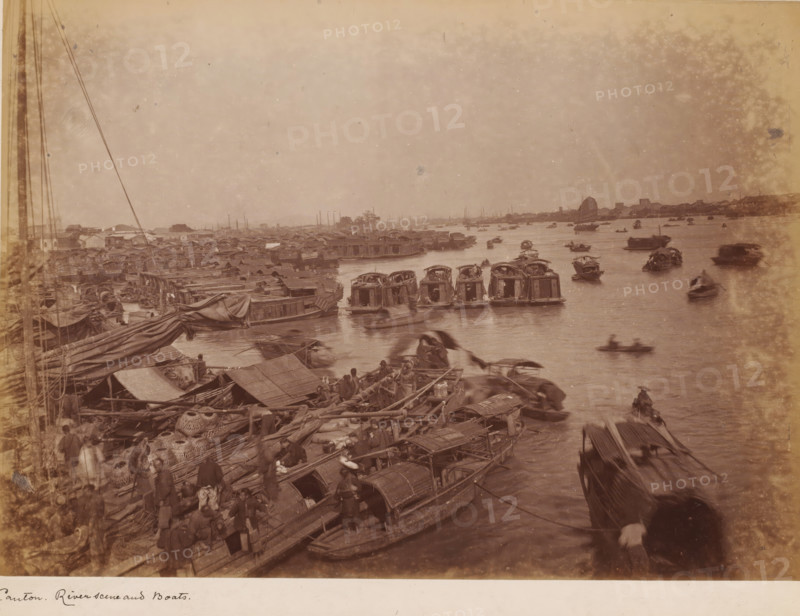 View of the river with boats near Guangzhou, photograph, annotation: 'Canton, Riverscene and boats', 1850 - 1900, photographer, paper, pasteboard, albumen print, height 230 mm x width 310 mm, harbour, river (landscape with figures, staffage), Canton (city), A wide river scene with many small wooden boats clustered near the bank and scattered across the water, several boats with curved canopies and others open, some boats docked alongside a crowded riverside area filled with tarps, woven baskets, ropes, and stacked cargo, numerous people standing or moving among the boats and along the dock, long masts and rigging visible on the left, a distant shoreline with low buildings and a single sail visible further out on the water, and a hazy sky above.