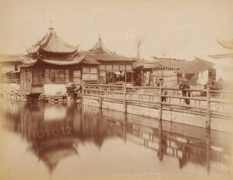 View of a temple the Shanghai, photograph, inscription: 'Sze Yuen Ming Co. 3 4, 42, Nanking Road', annotation: 'Shanghai. Chinese City', photographer: Sze Yuen Ming & Co, Shanghai, photographer: Sze Yuen Ming, 1850 - 1900, paper, pasteboard, albumen print, height 207 mm x width 258 mm, street (city(-scape) with figures, staffage), temple, shrine, traditional Chinese religions, A waterfront scene with ornate roofed buildings featuring curved eaves and tiled roofs, wooden framed windows, and lattice panels, a wooden railing running along a stone-lined water channel, reflections of the roofs and railing visible in the water, a few leafless trees rising above the rooftops, a sloped canopy supported by poles over a small area beside the railing, several people standing or leaning near the railing and under the canopy, simple wooden structures and fences behind the railing, stone and wooden elements at the water's edge, and muted sepia tones throughout the image.