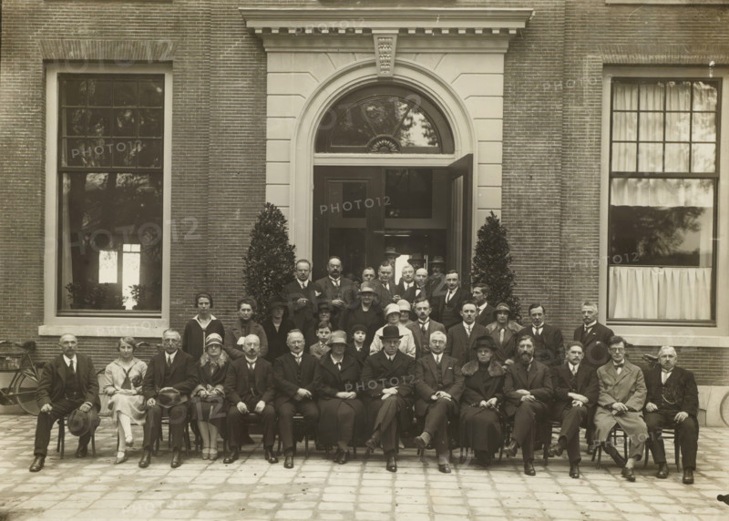 Group portrait of men and women in front of the entrance of a building, photograph, stamp: 'Copyright. Foto pers Bureau Hollandia Alkmaar, telephone 760', photographer: Hollandia, Foto Pers Bureau, Alkmaar, 1910 - 1930, paper, gelatin silver print, height 170 mm x width 235 mm, height 296 mm x width 355 mm, anonymous historical persons portrayed in a group, in a group-portrait, A large group of people arranged in rows in front of a brick building with an arched doorway and large rectangular windows, with two tall potted plants flanking the entrance; the men wear suits with ties or bow ties and many wear hats, some hold canes, and the women wear dresses or coats with cloche or brimmed hats, seated and standing on a paved courtyard with a bicycle visible at the left edge.