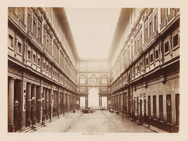 Gli Uffizi, Uffizi Gallery in Florence, Italy, Firenze, photograph, number: '3101.', address: 'GIACOMO BROGI, PHOTOGRAPHEDITORE, FIRENZE, LUNG ARNO DELLE GRAZIE 15', photographer: Giacomo Brogi, Florence, publisher: Edizioni Brogi, 1864 - 1881, Tuscany, pasteboard, paper, albumen print, height 345 mm x width 470 mm, permanent exhibition, museum, Uffizi Gallery, A long, narrow courtyard flanked by two symmetrical multi-story stone buildings with rows of tall, rectangular windows and decorative window frames; each building has a colonnaded ground level with evenly spaced columns and niches containing statues, ornate lamp posts along the walkways, and balustraded ledges beneath the upper windows; at the far end an arched gateway topped by a building section with large windows and sculptural reliefs; the paved courtyard shows scattered carts and a few indistinct objects near the central opening, and the entire scene appears in a sepia-toned photograph.