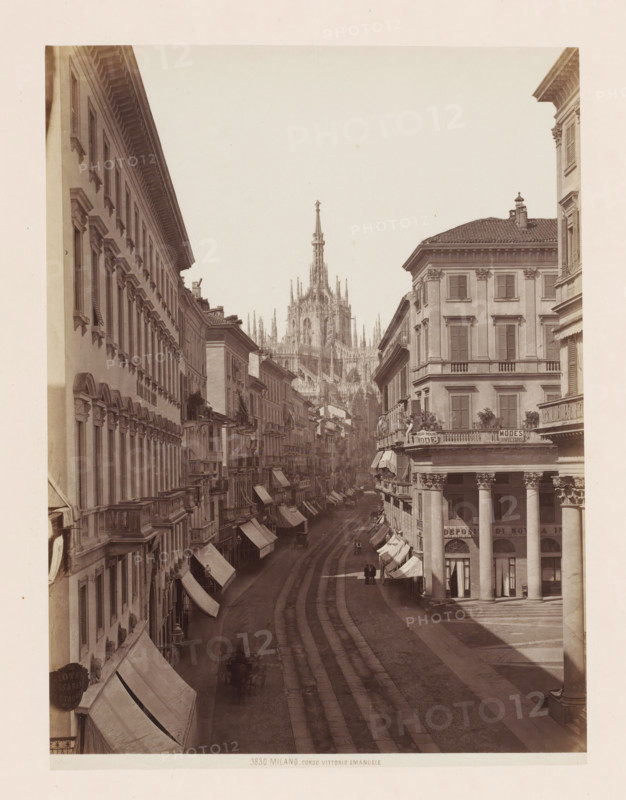 View of the Corso Vittorio Emanuele in Milan, Italy, Corso Vittorio Emanuele, Milano, photograph, number: '3830.', address: 'GIACOMO BROGI, FOTOGRAFO-EDITORE, FIRENZE, LUNG ARNO DELLE GRAZIE 15', photographer: Giacomo Brogi, Milan, publisher: Edizioni Brogi, Florence, 1864 - 1881, Tuscany, Lombardy, pasteboard, paper, albumen print, height 471 mm x width 352 mm, urban housing, street, Corso Vittorio Emanuele II, A wide street lined with tall multi-story buildings on both sides, many with shutters on the windows and decorative cornices, awnings extending over the sidewalks, and balconies visible on several facades; a grand cathedral with a pointed spire and many ornate pinnacles rises in the distance at the end of the street; the street surface shows parallel tracks and a few small figures walking or standing along the road, with some horse-drawn carriages and shadowed arcades beneath colonnades on the nearer corner building.
