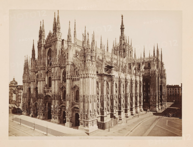 La Cattedrale dal Palazzo Reale, Exterior of the Cathedral of Milan, Italy, Milano, photograph, number: '3818.', address: 'GIACOMO BROGI, FOTOGRAFO EDITOR, FIRENZE, LUNG ARNO DELLE GRAZIE 15', photographer: Giacomo Brogi, Milan, publisher: Edizioni Brogi, Florence, 1864 - 1881, Tuscany, Lombardy, pasteboard, paper, albumen print, height 344 mm x width 470 mm, church (exterior), Cathedral (Milan), A large ornate Gothic cathedral fills the frame with a façade of pointed spires, pinnacles, and intricate stone carvings, including sculpted figures set into vertical niches. Tall arched doorways and large stained glass windows are visible amid clustered columns and buttresses. A central tower rises above the roofline with a slender spire topped by a statue. Steps and a paved area run along the front of the building, and a low fence lines the edge of the pavement. Additional buildings are visible at the edges of the scene.