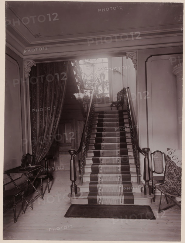 Stairwell in a house, photograph, address: '25 Drottninggatan 25, Göteborg.', photographer: Caroline Gaudard, Sweden, 1895 - 1897, paper, pasteboard, baryta paper, daylight collodion silver print, height 220 mm x width 163 mm, height 344 mm x width 282 mm, staircase, A grand wooden staircase with a patterned runner leads upward between turned newel posts and slender balusters, flanked by carved corner brackets and wall moldings; at the top of the stairs a decorative chair sits near a tall window through which large leafy plants are visible, and a pendant light hangs near the landing; to the left a heavy patterned drape partially covers a wooden door and a small table with folding legs and a stool, while to the right a cushioned chair with a patterned throw is positioned against the wall.