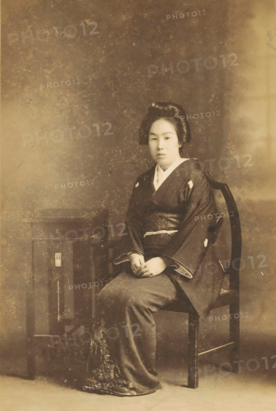 Portrait of an unknown Japanese woman, photograph, inscription: 'S. Hachiya. Kiriu. Gunma.', photographer: S Hachiya, Kiryu, c. 1890 - c. 1920, Gunma, paper, pasteboard, baryta paper, daylight gelatin silver print, height 137 mm x width 92 mm, height 228 mm x width 138 mm, adult woman, anonymous historical person depicted, A sepia-toned portrait of a seated woman wearing a traditional dark kimono with a lighter collar and patterned hem, her hands resting in her lap, hair styled in an updo, seated on a wooden chair next to a small wooden table, set against a mottled backdrop with visible age marks and surface wear.